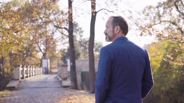 A Middleaged Caucasian Man Walks Toward a Bridge and Smiles Over His Shoulder at the Camera