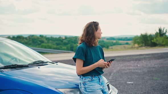 Young Woman Uses Her Phone Leaning on the Bumper of a Car and Enjoying the View