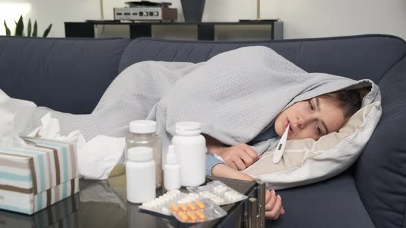 Sick Young Woman with Fever Lying on Sofa Covered in Blanket Having Flu Symptoms Holding and Looking