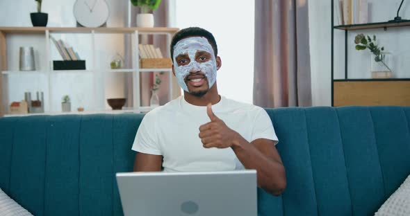 African American with Facial Mask Sitting on the Sofa with Computer and Gesturing Thumb Up