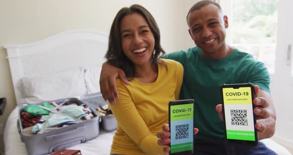 Happy biracial couple sitting on bed and showing smartphones with covid 19 passports on screens