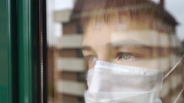 Young man in quarantine close-up. In the face medical mask looks out the window with sad eyes.