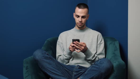 Young Caucasian Man Uses Smartphone While Sitting on a Chair at Home. Man Browses Through Internet