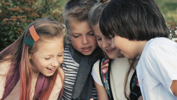 Four happy children sit outdoors and use mobile phone smiling.