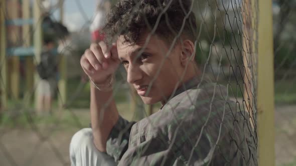 Side View of Unhappy African American Young Man Sitting in Sunlight on Outdoor Sport Court As