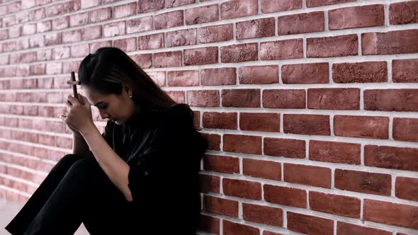 Christian Asian woman is sitting against a wall, feeling sad, holding a cross and praying to God.