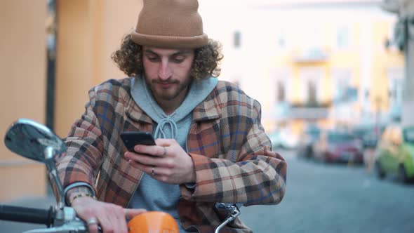 Cheerful curly-haired man texting by phone while sitting on moped