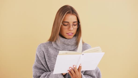 Young Lady in Glasses is Reading a Book and Smiling While Posing Against Beige Studio Background