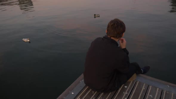 Overhead view of a young man sitting on the edge of a lake pier eating potatoe chips and looking at