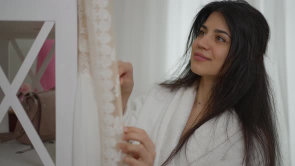 Beautiful Smiling Happy Middle Eastern Bride Admiring Wedding Dress Hanging in Living Room