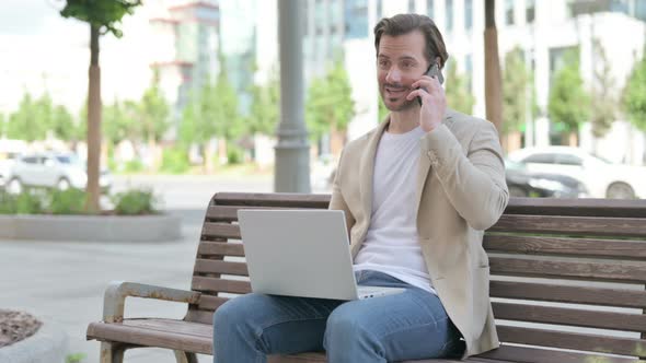 Young Man Talking on Phone and Using Laptop While Sitting on Bench