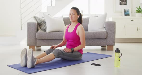 Asian woman on mat wearing earphones, exercising with smartphone at home