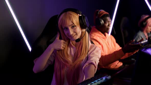 Teenage Gamer Girl Wearing Headset Holding Up Colorful Keyboard and Giving Thumbs Up After Winning