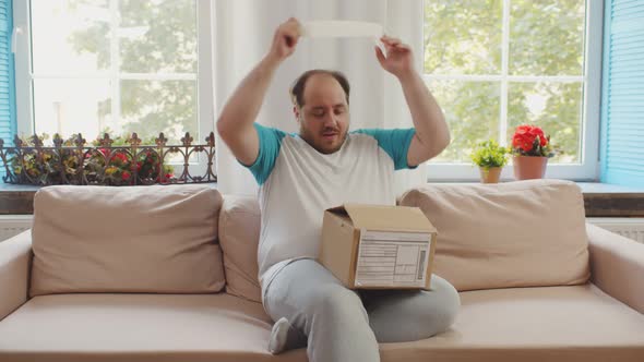 Happy Stout Man Opening Parcel with Blue Dumbbells Ordered Online Sitting on Couch at Home