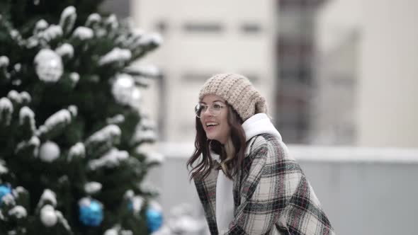A Pretty Young Darkhaired Woman in Round Glasses and a Knitted Hat is on a City Street in the