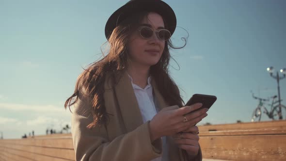 Brunette Girl Is Using Smartphone, Sitting Outdoors in Sunny Day, People Behind