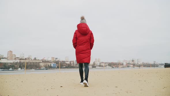 A young girl warmly dressed in a red down jacket and gray hat is walking along the river