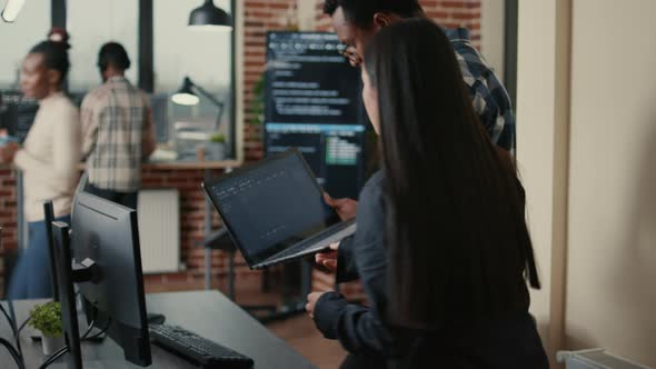 Two Programers Holding Laptop with Coding Interface Walking Towards Desk and Sitting Down