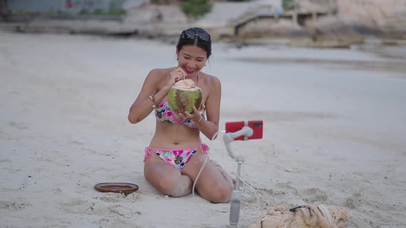 Asian Thai Girl Recording Live Stream Sitting on a Beach and Drinking Coconut Water at Sunset