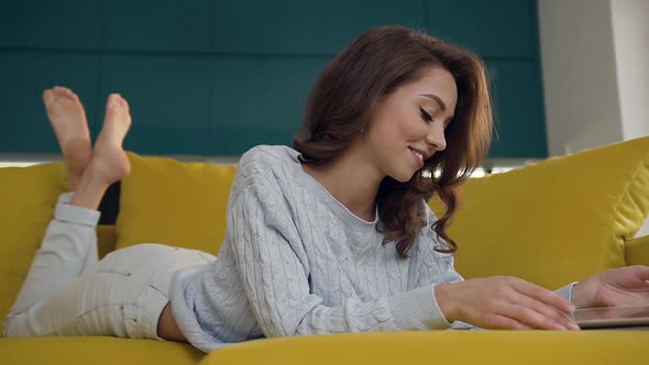 Young Woman with Wavy Hair Laying on the Cozy Sofa and Using Tablet PC