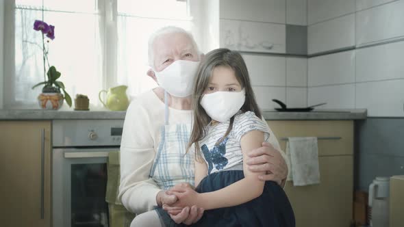 Grandmother Hugs Her Granddaughter Sitting In The Kitchen. They Are Both In Medical Masks