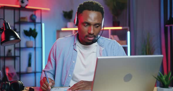 African American in Headset Writing Down Important Notes Into Report During Video Meeting