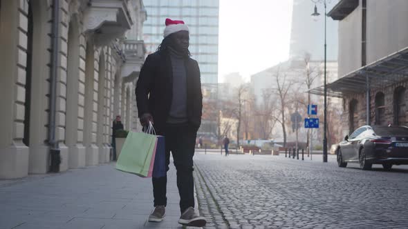 Black Man with Christmas Hat Walking on the City Street with Christmas Gifts