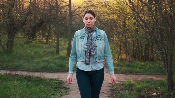 A Beautiful Brunette Girl in a Denim Jacket and Scarf Walks in the Park Among the Trees