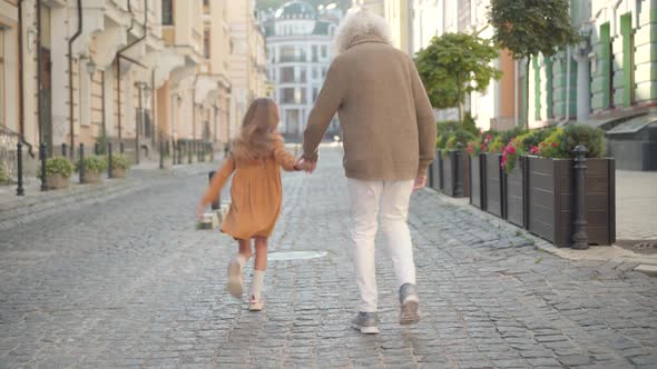 Wide Shot Back View of Joyful Little Caucasian Girl Jumping As Walking with Senior Grey-haired Man