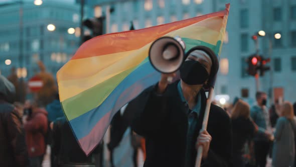 Woman with Face Mask Speaking Into the Megaphone While Holding Rainbow Flag. Demonstration Against