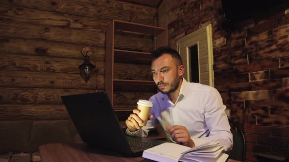 A Businessman is Drinking Coffee Looking at the Screen and in a Notebook