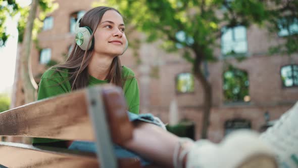 Positive blonde woman wearing green t-shirt listening music in headphones on the bench