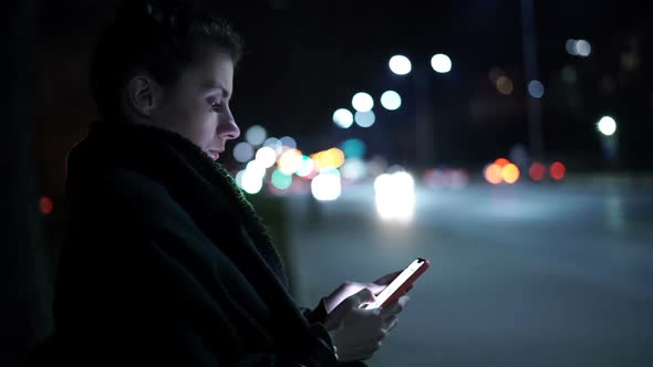 Side View of Young Woman Using Smartphone with Night City on Background