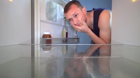 man looks into an empty refrigerator and takes out bottle of alcohol. Alcoholism