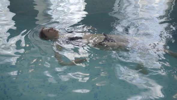 Top View of Young Caucasian Woman Swimming on Back Along Indoor Basin. Wide Shot of Slim Beautiful