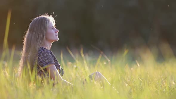 Young Woman with Long Hair Sitting Outdoors in Summer Field Grass Enjoying Nature at Sunset