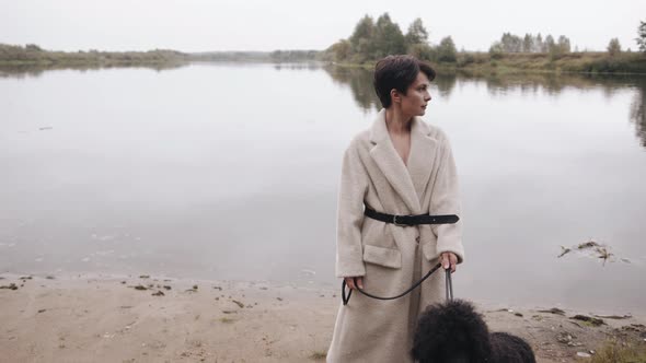 An Exquisite Young Woman with Short Hair Walks Along the River Bank with a Poodle on a Leash and