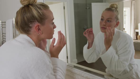 Caucasian woman in bathrobe touching her face while looking in the mirror in the bathroom