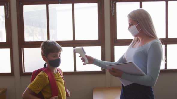 Female teacher wearing a face mask measuring temperature of a boy in class at school