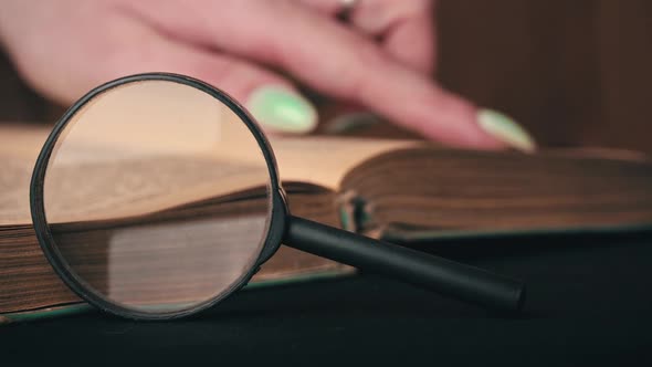Girl Turns the Page of an Old Book Next to a Magnifying Glass