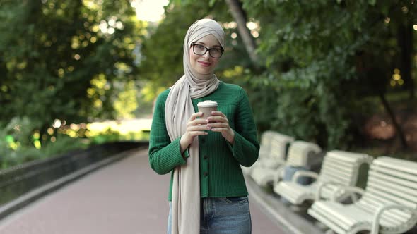 Pretty Smiling Muslim Woman in Hijab Walks at Green Summer City Park with Coffee to Go