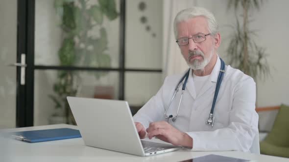 Senior Old Doctor Looking at Camera While Using Laptop in Clinic