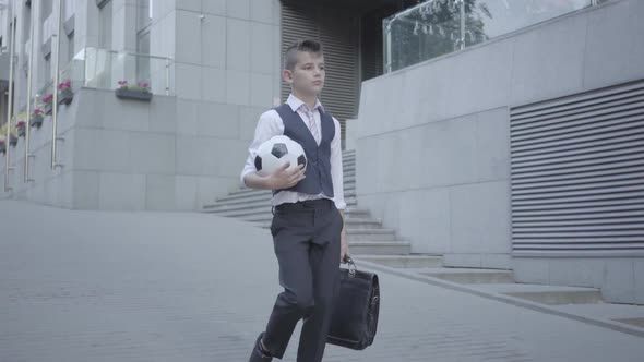 Serious Well-dressed Boy Walking Down the Street Holding the Soccer Ball and Purse in Hands