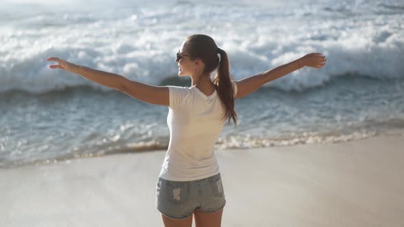 Backside View of Woman Looks at Ocean on Beach and Spreads Arms Wide Open