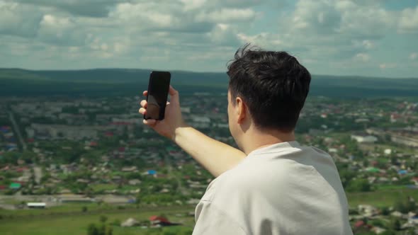 A Young Adult Male Takes a Smartphone Picture From the Top of a Large Hill