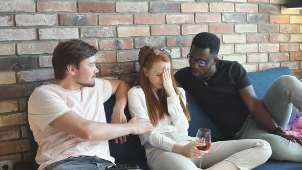 Redhead Woman Crying Sitting on the Bed with a Glass of Champagne Among Two Different Men.