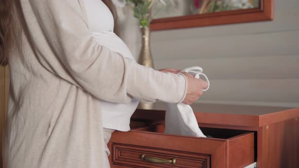 Cute Happy Pregnant Woman Pulls Out Baby Clothes From Dresser Touching Her Tummy at Home