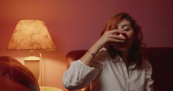 Young Italian girl with white shirt drinks coffee sitting on the sofa at home