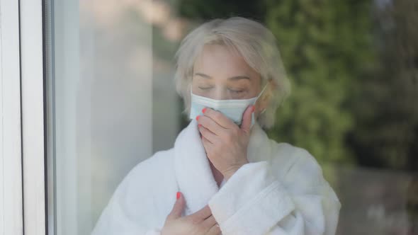 Ill Woman in Coronavirus Face Mask Coughing and Sneezing Standing at Home Looking Out Window