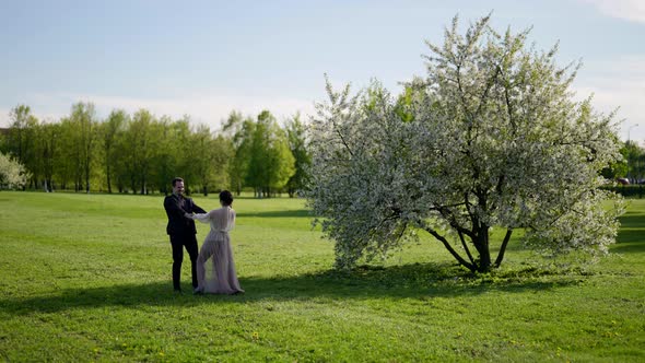 Man and Woman are Holding Hands and Whirling on Lawn Near Blooming Cherry Tree in Garden